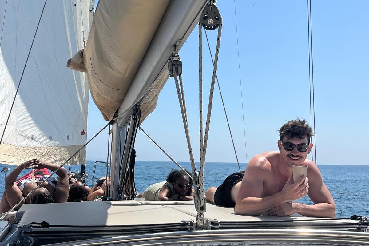 Charlie lying on the deck of a sailing boat in sunglasses, with the sail up and open blue sea behind him.