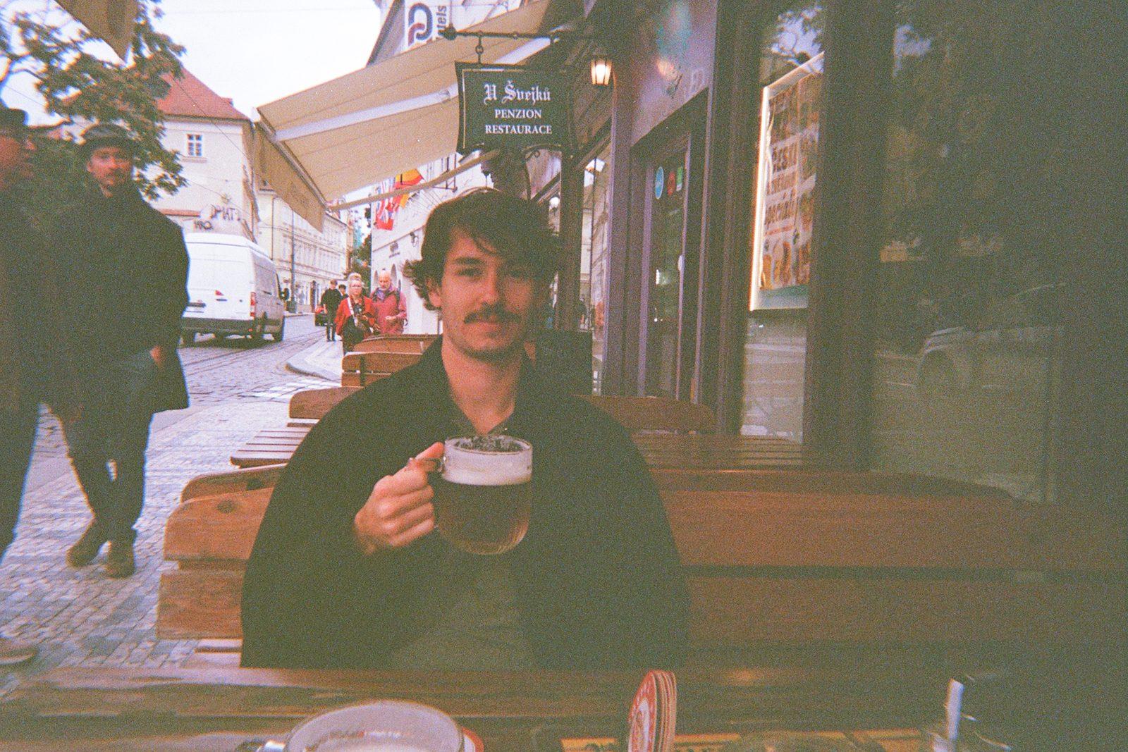 A film photograph of Charlie sitting at an outdoor table in Prague in a dark jacket, holding a glass of beer, with a sign reading U Švejků on the building behind him.
