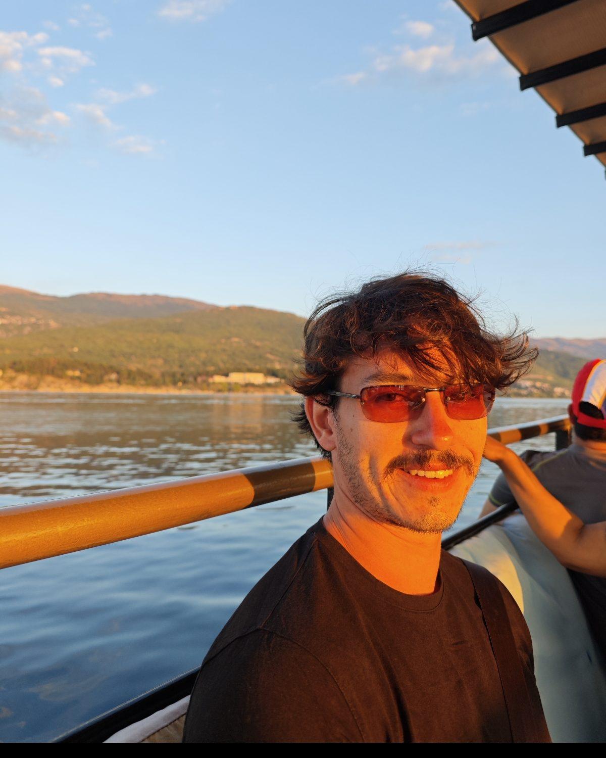 Charlie on a boat at sunset in a black t-shirt and red-tinted sunglasses, smiling at the camera, with a yellow rail and hills across the water behind him.